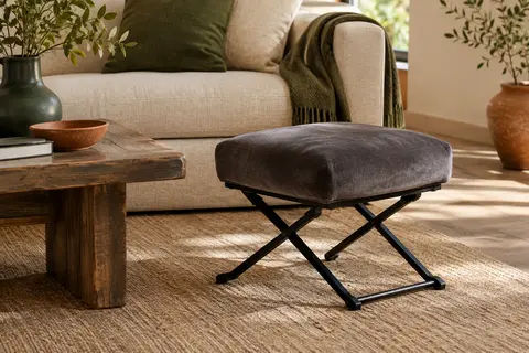 Repose-pieds pliable en velours gris foncé avec pieds métalliques en X dans un salon lumineux.