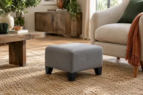Pouf repose-pieds compact gris à coussin rembourré et pieds noirs, installé près d’un fauteuil dans un salon lumineux