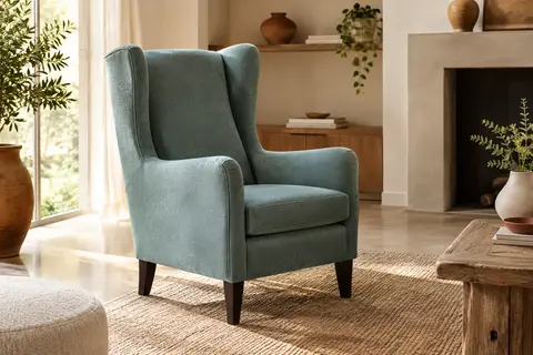 Fauteuil à oreilles Carla en tissu bleu vert d’eau, avec dossier haut et pieds couleur wengé dans un salon lumineux.