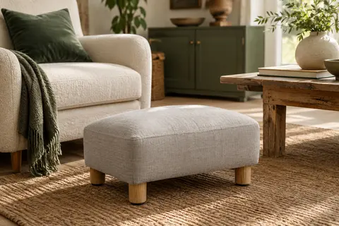 Repose-pieds ottoman gris clair rembourré, avec pieds en bois, près d’un fauteuil dans un salon lumineux.